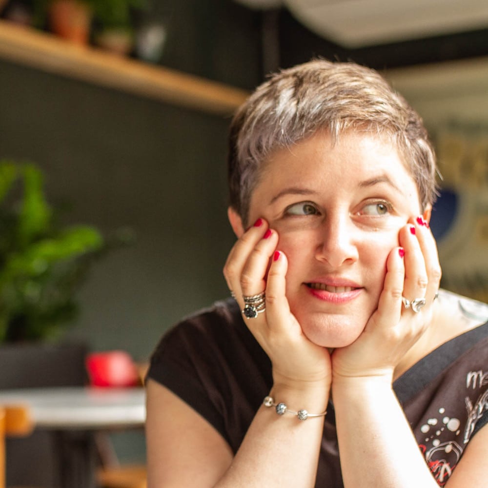 Image of trauma recovery support coach Emily, a middle-aged white woman with cropped hair. She is sitting in a café with her chin resting on her hands. She is looking off to side and smiling