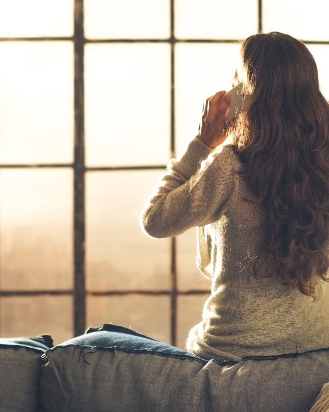 Image of a white brunette woman from behind. She is leaning against a sofa looking outwards & she is on the phone.