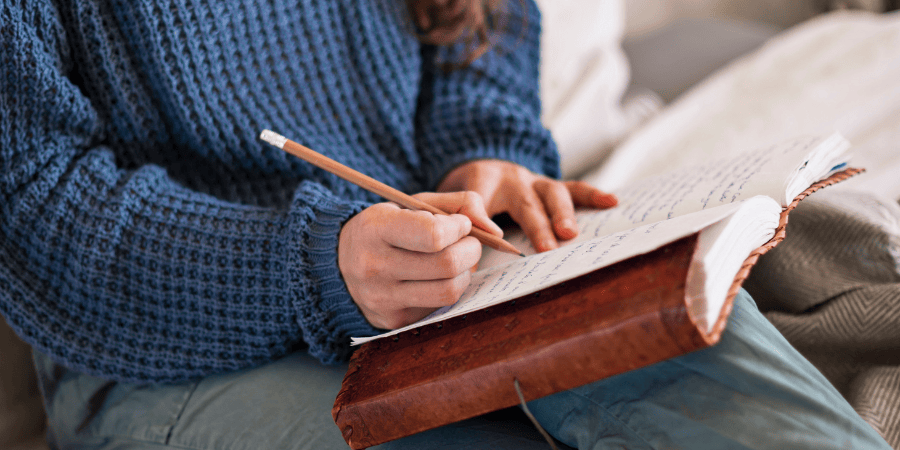 Image of a white brunette woman up close on her torso & lap. She is writing in a notebook/journal. It gives the impressions she is working through something on a path to recovery. It gives the impression of someone working through their trauma and going on a journey of recovery, rediscovery and reconnection.