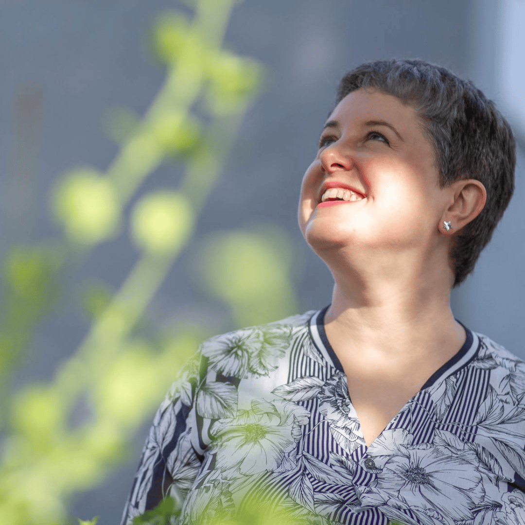 Image of Emily, a middle-aged white woman with cropped hair. Emily is outside & looking away from the camera. Her face is dappled by the sun and there is green foliage in the foreground. She looks happy & at peace & able to coach you after trauma