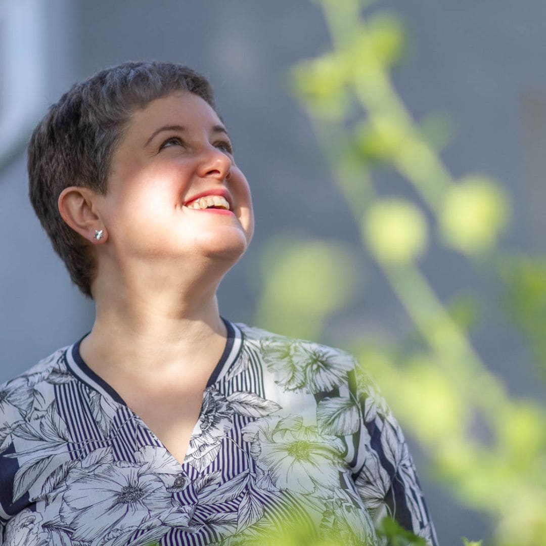 Image of trauma recovery support coach Emily, a middle-aged white woman with cropped hair. Emily is outside & looking away from the camera. Her face is dappled by the sun and there is green foliage in the foreground. She looks happy & at peace.