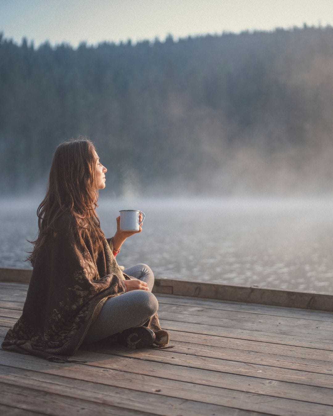 White brunette woman sitting on the end of a wooden pier, with a mug in her hand looking over the water.