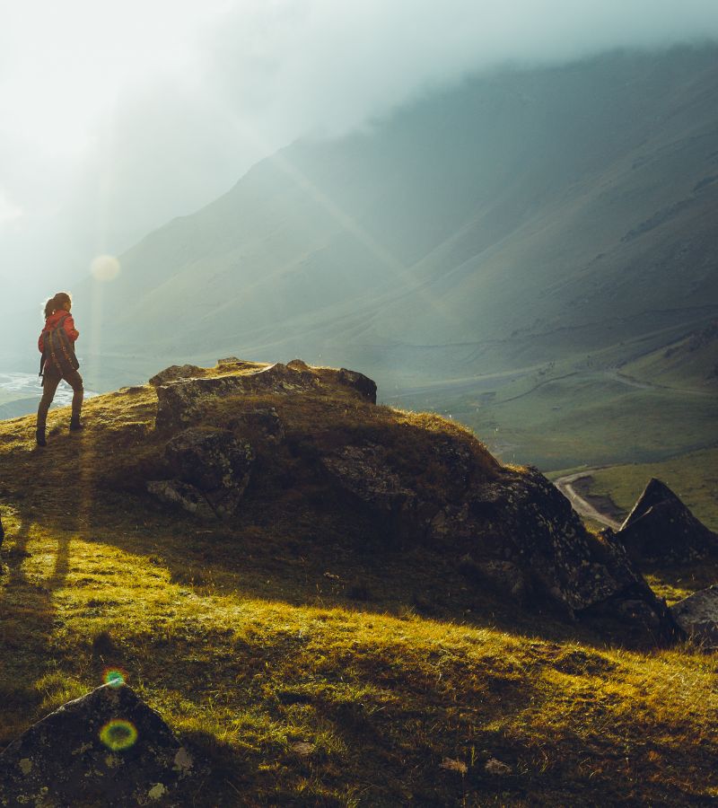 Image of a woman at the top of one mountain looking out onto the view & more mountains. It is a very serene & special feel. It depicts the feeling of having gone on a trauma recovery journey.