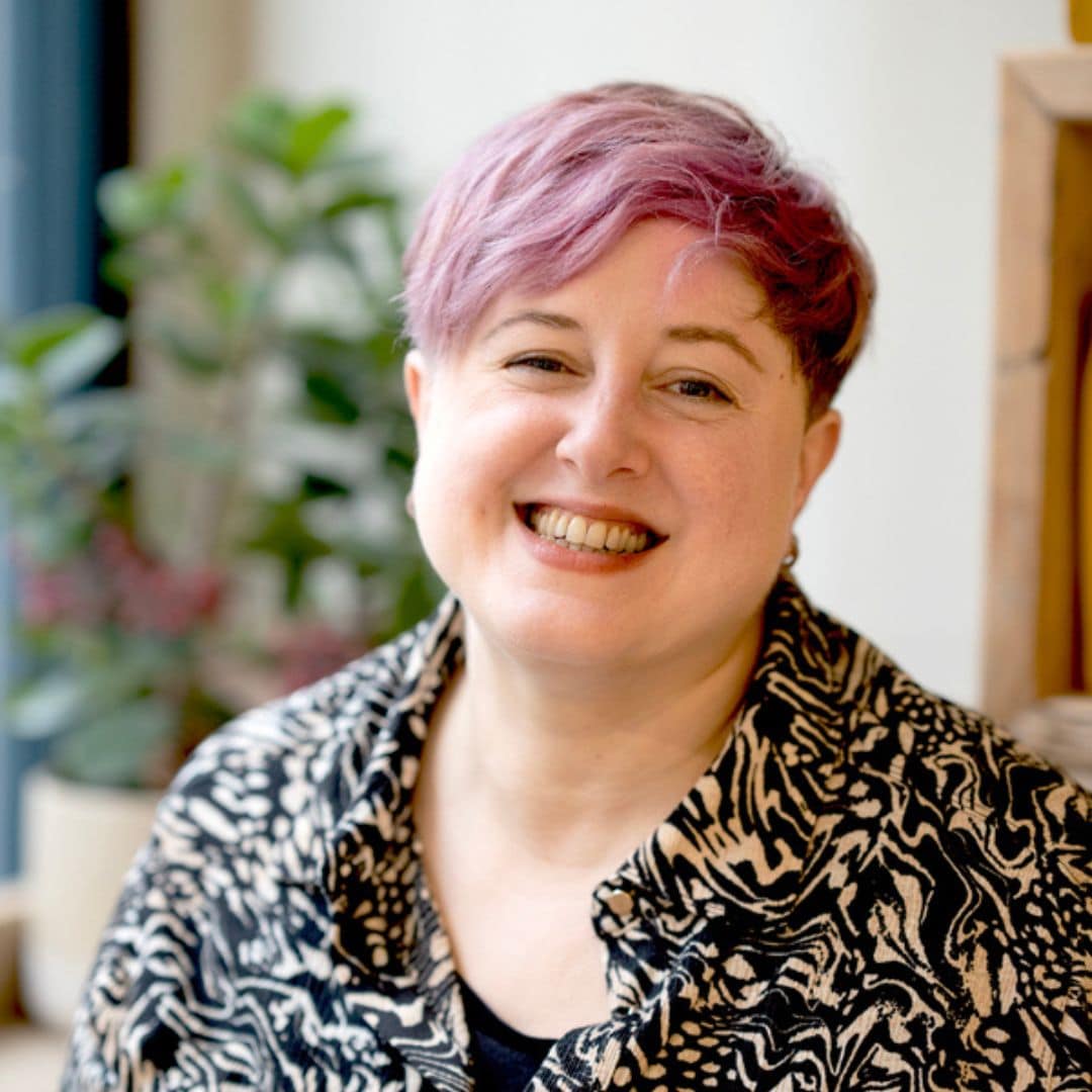 Image of trauma recovery support coach Emily, a middle-aged white woman with cropped hair. Emily is outside & looking away from the camera. Her face is dappled by the sun and there is green foliage in the foreground. She looks happy & at peace.