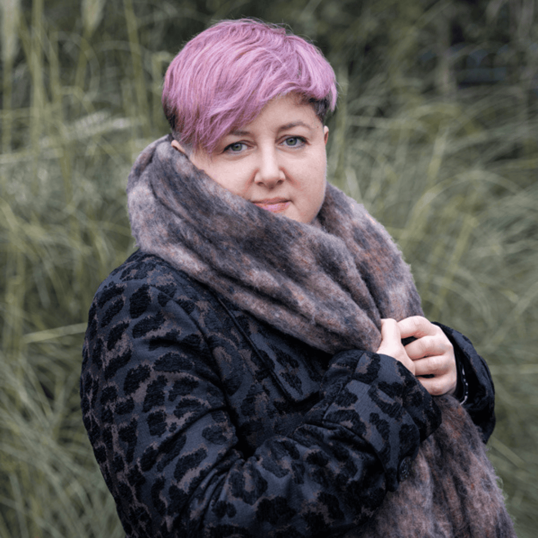 Image of trauma recovery support coach Emily, a middle-aged white woman with cropped hair. She is sitting in a café with her chin resting on her hands. She is looking off to side and smiling