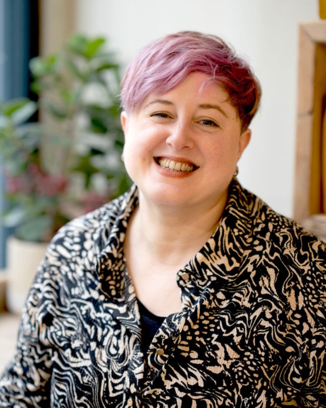 Image of Emily, a middle-aged white woman with cropped hair. Emily is outside & looking away from the camera. Her face is dappled by the sun and there is green foliage in the foreground. She looks happy & at peace & able to coach you after trauma