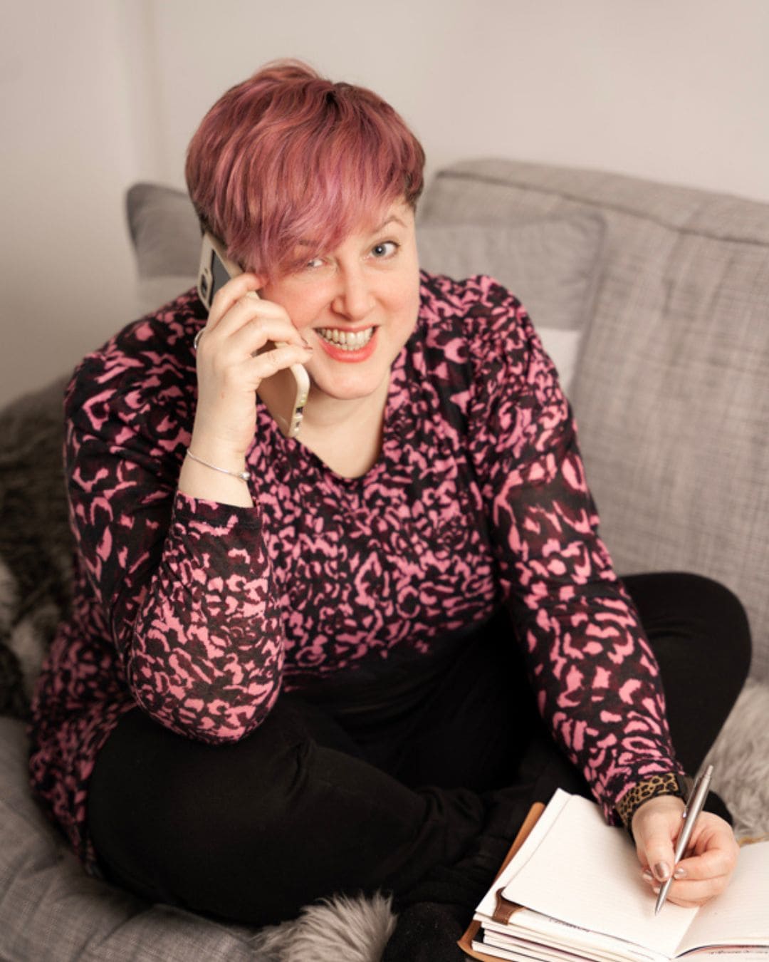 Image of Emily sitting cross legged on a sofa, she is on the phone and poised as if about to write something in her notebook. She is looking at the camera & smiling.